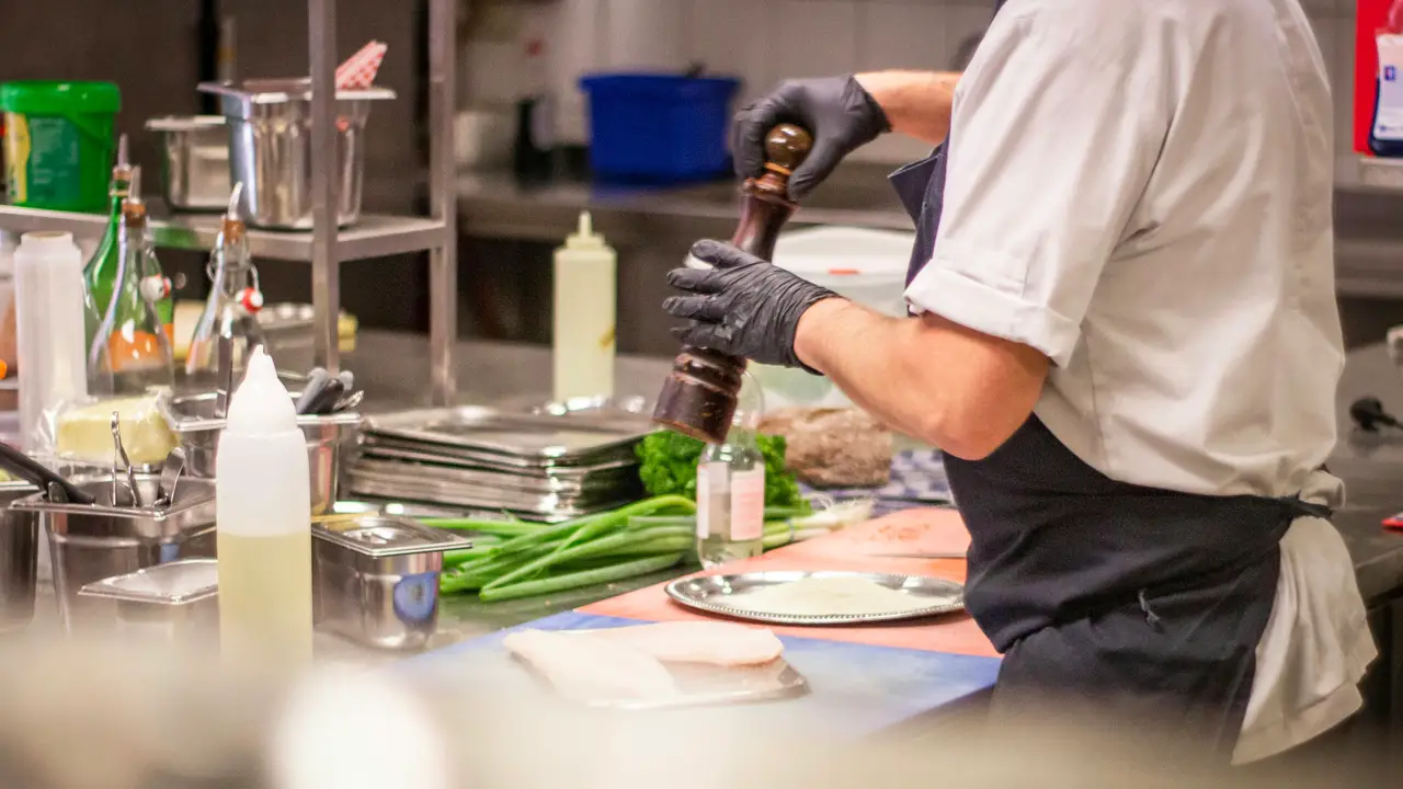 Cuisinier ganté qui moud du poivre au-dessus d’un plat, sur un plan de travail en inox, entre bacs gastro et jeunes oignons